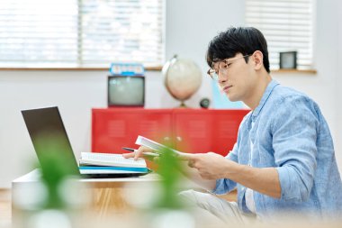 Genç Asyalı Adam Parlak Modern Oturma Odasında Laptop ve Kitapla Çalışıyor