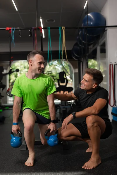 Two men, coach and trainee look at each other smiling while doing exercise at the gym