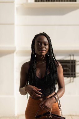 Genç siyah kadın çantasını tutarken şık poz veriyor ve gün batımında sokakta kameraya bakıyor.