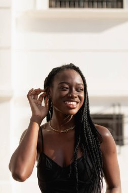 Genç siyah bir kadın elleriyle saçlarını düzeltirken mutlu bir şekilde gülümsüyor.