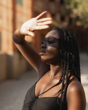 Saçında örgüler olan genç bir zenci kadın şehirde yürürken elleriyle gözlerini güneşten koruyor.