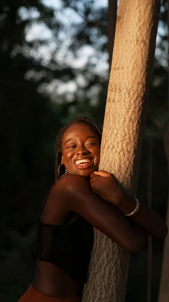 一位留着辫子的年轻黑人妇女在公园里散步时高兴地抱着一棵树 — 图库照片