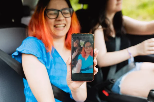 A young woman laughs happily, while showing a selfie photo of her and her friend, that she has taken with the smartphone, inside the car