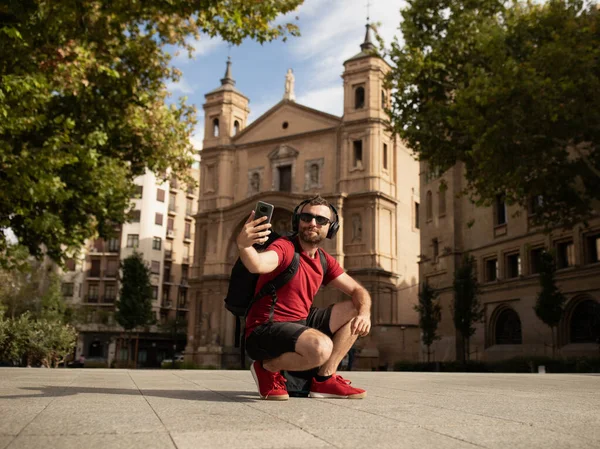 一位年轻的男性旅行者和背包客 戴着太阳镜 对着摄像机笑着 一边拿着手机在西班牙一个城市观光 — 图库照片