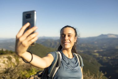 Genç, mutlu bir kadın dağın tepesinden akıllı telefonuyla selfie çekerken gülümsüyor.