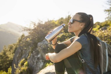 Genç bir kadın, bir dağcı, bir dağın tepesinde otururken su içer, arka planda odak dışı doğal manzara