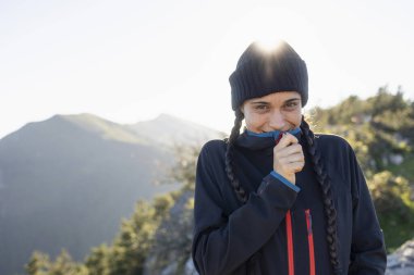Genç bir kadın, dağcı, dağın tepesinde soğuktan korunan, gülümseyen ve manzaranın ve doğanın tadını çıkaran.