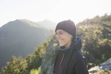 Genç bir kadın, dağcı, dağın tepesinde soğuktan korunan, gülümseyen ve manzaranın ve doğanın tadını çıkaran.