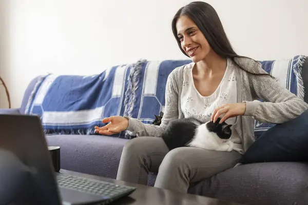 A young woman chats with someone on a video call via a laptop computer while petting her cat, sitting on the sofa at home.
