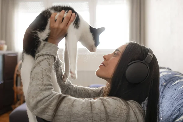 A woman listens to music, with headphones, sitting on the sofa at home, while playing with and cuddling her cat