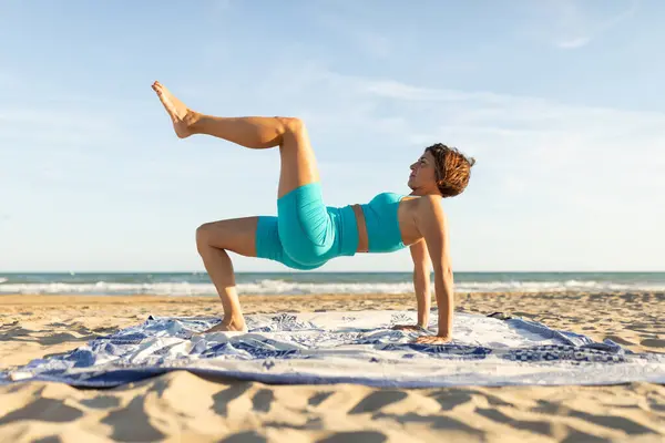 Bir kadın plajda pilates egzersizi yapıyor ve bacak çekme hareketi yapıyor.