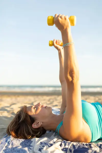 一名妇女躺在海滩上 用轻便的1公斤哑铃进行伸展和放松运动 图库图片