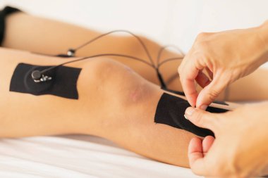 Bir fizyoterapist elektrostimülasyon tedavisi sırasında dizindeki spor yaralanmasından dolayı elektrotları genç bir hastanın bacağına yerleştirir.