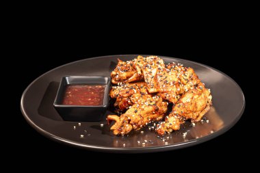 Acı soslu tavuk kanadı ızgarada kızartılmış susam tohumları serpiştirilmiş. Yakınlarda sıcak ketçap var. Karanlık bir arka planda.