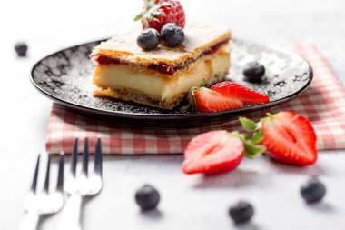 Bir parça Napolyon pastası haşhaş tohumu ve kirazla süslenmiş, çilekle süslenmiş, hafif arkaplanlı yaban mersini. Kapat.