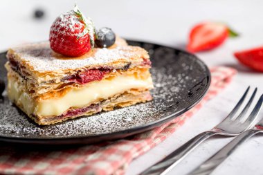 Bir parça Napolyon pastası haşhaş tohumu ve kirazla süslenmiş, çilekle süslenmiş, hafif arkaplanlı yaban mersini. Kapat.