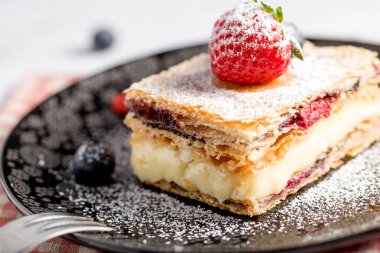 Bir parça Napolyon pastası haşhaş tohumu ve kirazla süslenmiş, çilekle süslenmiş, hafif arkaplanlı yaban mersini. Kapat.