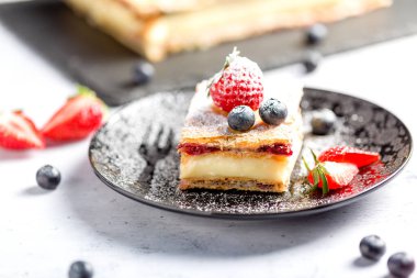 Bir parça Napolyon pastası haşhaş tohumu ve kirazla süslenmiş, çilekle süslenmiş, hafif arkaplanlı yaban mersini. Kapat.