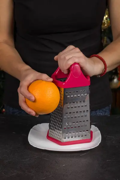 The hand of a Caucasian woman is rubbing an orange on a metal grater. This zest will be used to make the cake dough.