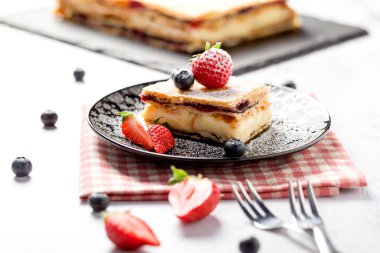 Bir parça Napolyon pastası haşhaş tohumu ve kirazla süslenmiş, çilekle süslenmiş, hafif arkaplanlı yaban mersini. Kapat.