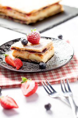 Bir parça Napolyon pastası haşhaş tohumu ve kirazla süslenmiş, çilekle süslenmiş, hafif arkaplanlı yaban mersini. Kapat.