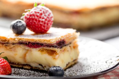 Bir parça Napolyon pastası haşhaş tohumu ve kirazla süslenmiş, çilekle süslenmiş, hafif arkaplanlı yaban mersini. Kapat.