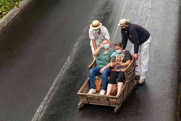 stock image FUNCHAL, PORTUGAL - AUGUST 24, 2021: Unidentified women ride happily down the street in wicker baskets (sleds) with the help of drivers in traditional uniforms.