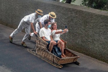 FUNCHAL, PORTUGAL - 24 AĞUSTOS 2021: Kimliği belirsiz genç çift geleneksel üniformalı sürücülerin yardımıyla sepetlerle (kızakçılar) caddede yuvarlanıyor.