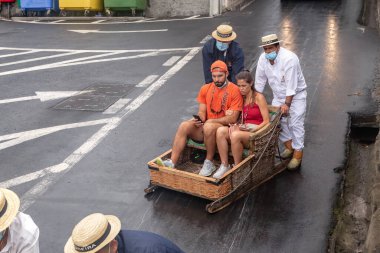 FUNCHAL, PORTUGAL - 24 AĞUSTOS 2021: Kimliği belirsiz genç çift geleneksel üniformalı sürücülerin yardımıyla sepetlerle (kızakçılar) caddede yuvarlanıyor.