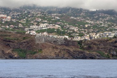 MADEIRA ADASI, PORTUGAL - 25 Ağustos 2021: Bu, adanın dağlık yamaçlarında yerleşim gelişimi olan okyanustan kayalık bir kıyıya bir manzara..
