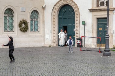 ROMA, İTALYA 8 MARTH 2023: Bir rahip ve bir cemaat eski şehrin merkezindeki bir meydanda, bir kilisenin duvarlarında oynayan çocukları izliyor..