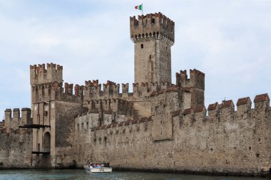 SIRMIONE, ITALY - MAY 13, 2018: A boat with unidentified tourists is located near the walls of the medieval Scaliger Castle on Lake Garda. clipart