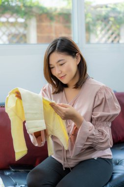 Doğum kıyafetleri içinde mutlu, genç, pragnent Asyalı bir kadın. Gülümsüyor ve elinde tuttuğu yeni bebek kıyafetlerine bakıyor. Parlak evinin oturma odasında otururken.