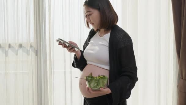 Счастливая Беременная Азиатка Пользуясь Смартфоном Ест Свой Здоровый Салат Гостиной — стоковое видео