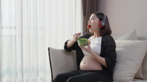 Szczęśliwa Azjatka Ciąży Jedząca Zdrową Sałatkę Słuchając Muzyki Słuchawki Salonie — Wideo stockowe