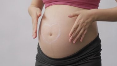 Hamile bir kadının göbeğini ovuşturup masaj yaparken, göbek deliğinin etrafında kalp şeklinde gerilme önleyici krem sürerken, yavaş çekimde vurulurken.