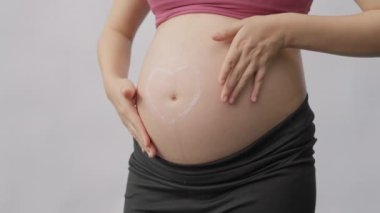 Hamile bir kadının göbeğini ovuşturup masaj yaparken, göbek deliğinin etrafında kalp şeklinde gerilme önleyici krem sürerken, yavaş çekimde vurulurken.