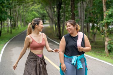Sıska ve kilolu iki genç kadın, yerel bir parkın koşu pistinde sabah yürüyüşü yaparken birbirlerine eşlik etmekten zevk alırlar.