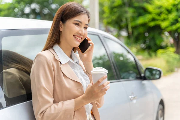 快乐微笑的年轻职业女性一边拿着一杯咖啡一边打电话 靠在自己的车上 图库图片