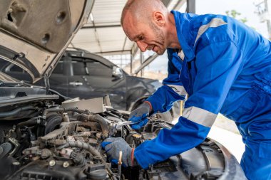 Mavi tulumlu bir erkek araba tamircisi araba tamirhanesindeki motoru tamir etmek için çok çalışıyor.