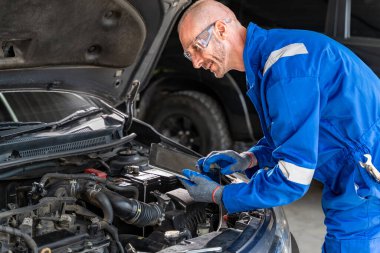 Erkek bir araba tamircisi arabasının tamirhanesinde çalışırken bilgisayarını kullanarak aracın motorunu kontrol ediyor.