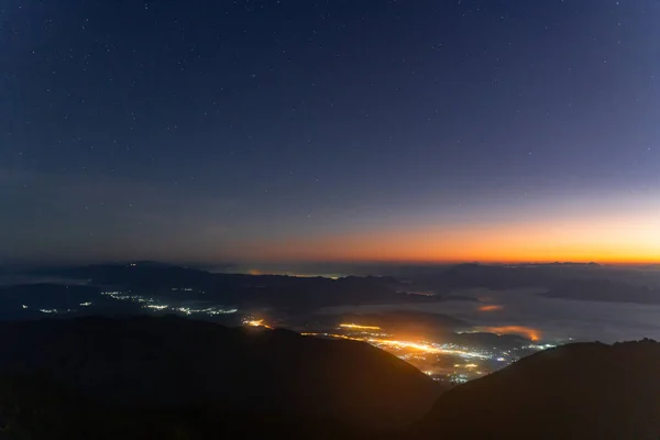 Early morning night sky from the top of Doi Luang Chiang Dao in Chiang Mai, Thailand overlooking a glowing town lights and beautiful dark blue sky with stars and glowing golden sun light at the horizon