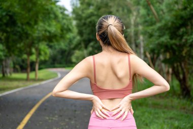 Sırtı ağrıyan uzun saçlı bir kadının arka planı. Ellerini beline koyuyor. Yerel bir parkın koşu yolunda dikilirken belini indiriyor.