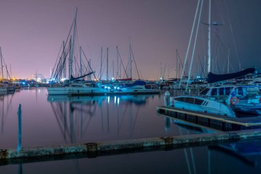 Sakin sularla dolu bir tekne rıhtımının gece görüşü ve arka planda birçok tekne ve uzak şehir ışıkları.