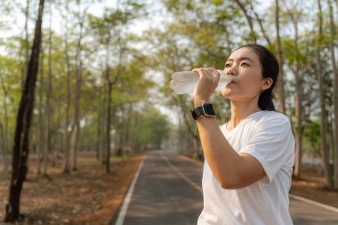 Genç bir kadın, sabah koşusundan sonra yerel bir parkta plastik şişeden su içiyor.