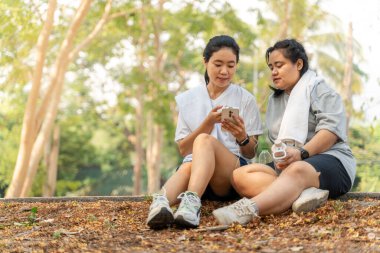 İki mutlu Asyalı bayan arkadaş sabah koşularından sonra kıvrımlı yolda birlikte otururken telefondan müzik dinliyorlar.