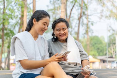 İki mutlu Asyalı bayan arkadaş sabah koşularından sonra kıvrımlı yolda birlikte otururken telefondan müzik dinliyorlar.