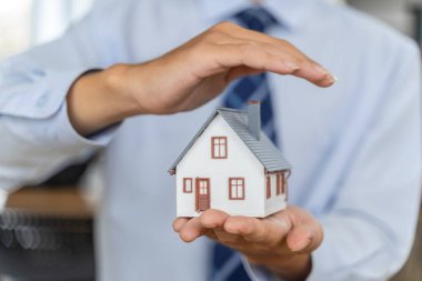 Erkek bir sigorta acentesinin bir elinde küçük bir ev modelini, diğer elinde ise ev sigortası konseptini korumak için tutarken ellerini kapat.