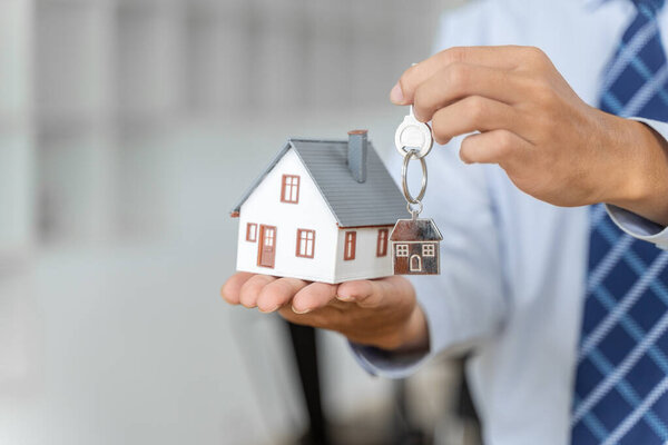 Close up hands of a realtor holding house key chaing and a small house model, buying home or real estate concept