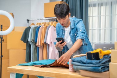 Genç online satıcı telefonunu gömlek ürününün fotoğraflarını çekmek için kullanıyor. Ev stüdyosundaki müşterilerine göndermek için.
